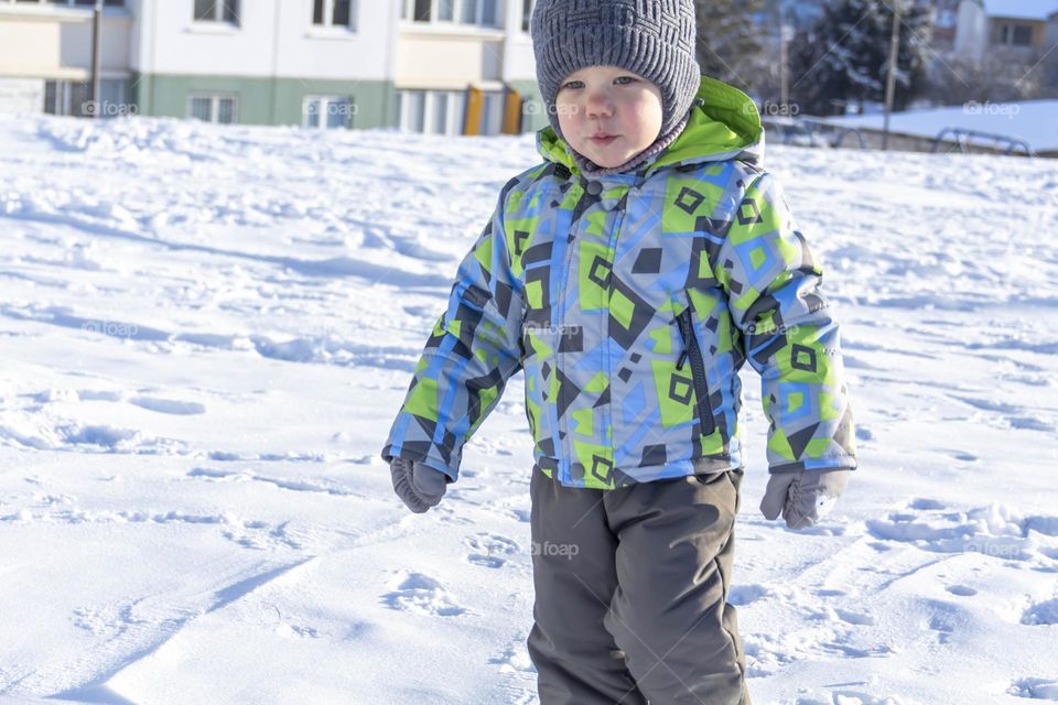 A child in winter in winter jackets, pants, a hat and boots on white snow on the street and in the park in nature plays winter fun and sleds.