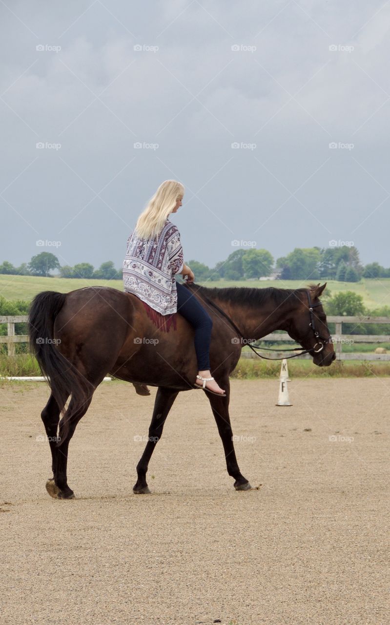 Blonde girl riding a dark bay horse bareback with an English bridle in a sandy arena. 
