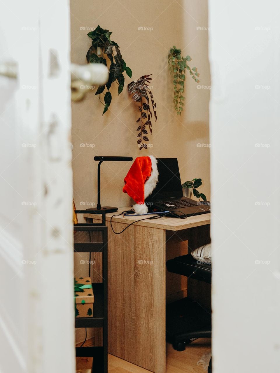 Beautiful view through open door to desktop with laptop in santa claus hat and hanging flowers on bedroom wall during daytime,close up view from side.
