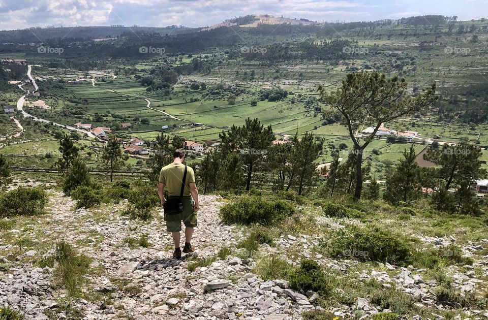 A man walks along a stony trail amidst green rolling hills and trees 