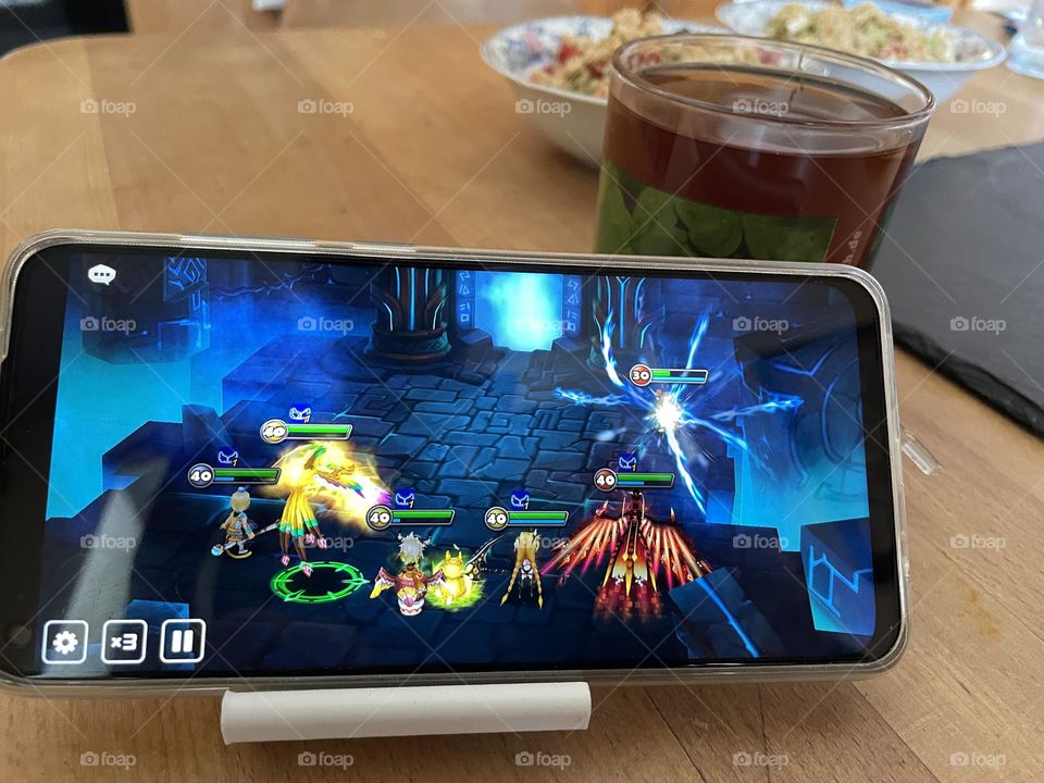 On a table with food and drinks there is a mobile phone playing a video game 