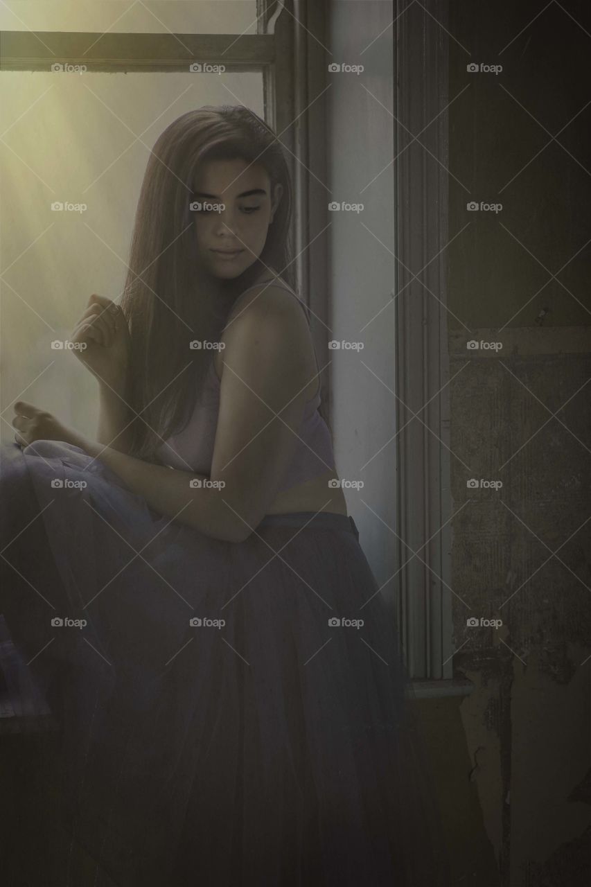 Girl seated on a window sill in a derelict building. She is looking down as if thinking or contemplating. There is a texture added to the photograph