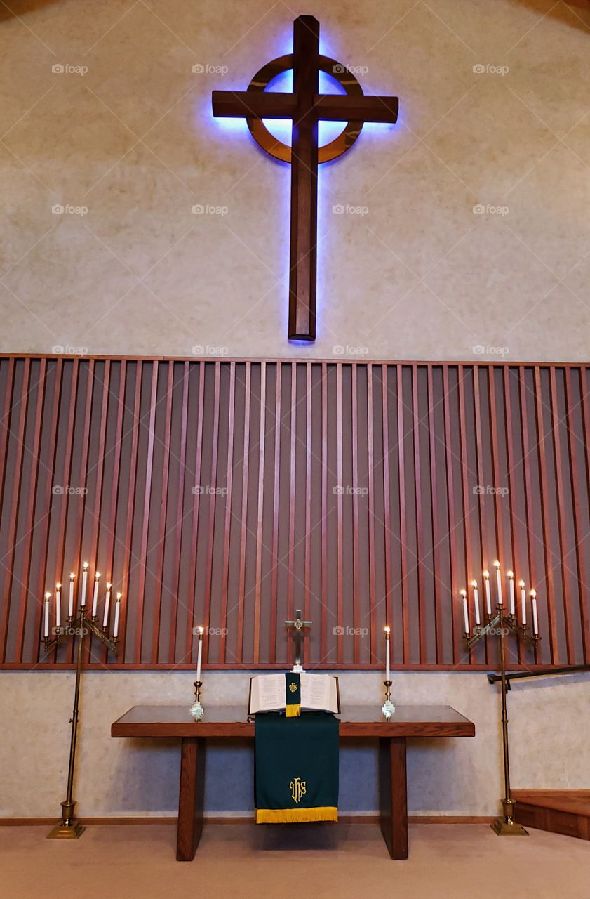 An alter setting with a beautiful table with candles on the side, a large open bible on a stand, and green and yellow paraments draped from the table with a large wooden cross above with a purple glow.