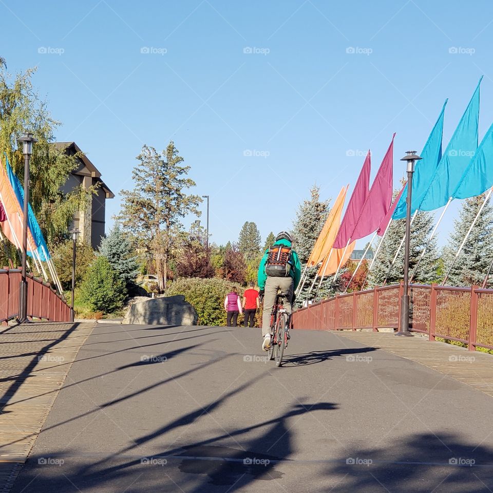 A couple walks for morning exercise while a bicyclist rides on his morning commute across a bridge adorned with colorful flags at the Old Mill District in Bend in Central Oregon on a sunny fall day. 
