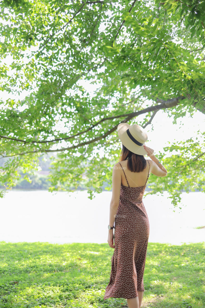 Summer weekend, nature landscape with girl portrait from back view. Young woman in a polka dot dress and a straw hat standing back against a background of green trees.