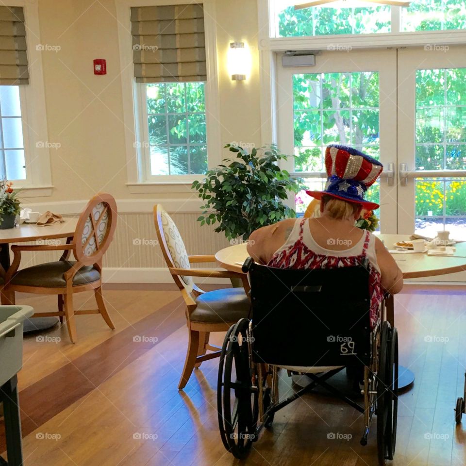 Nursing home patient sitting in wheelchair wearing American Flag 🇺🇸 hat celebrating July 4th Independence Day.