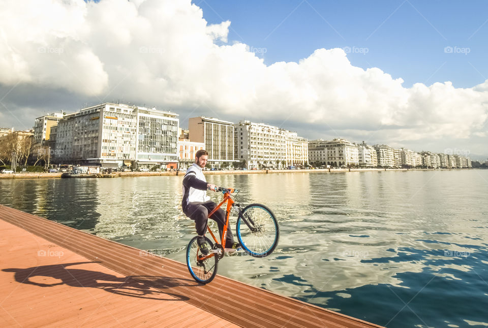 Teenager Boy Performing A Wheelie With His Bike In Front Of The Sea At The Dock
