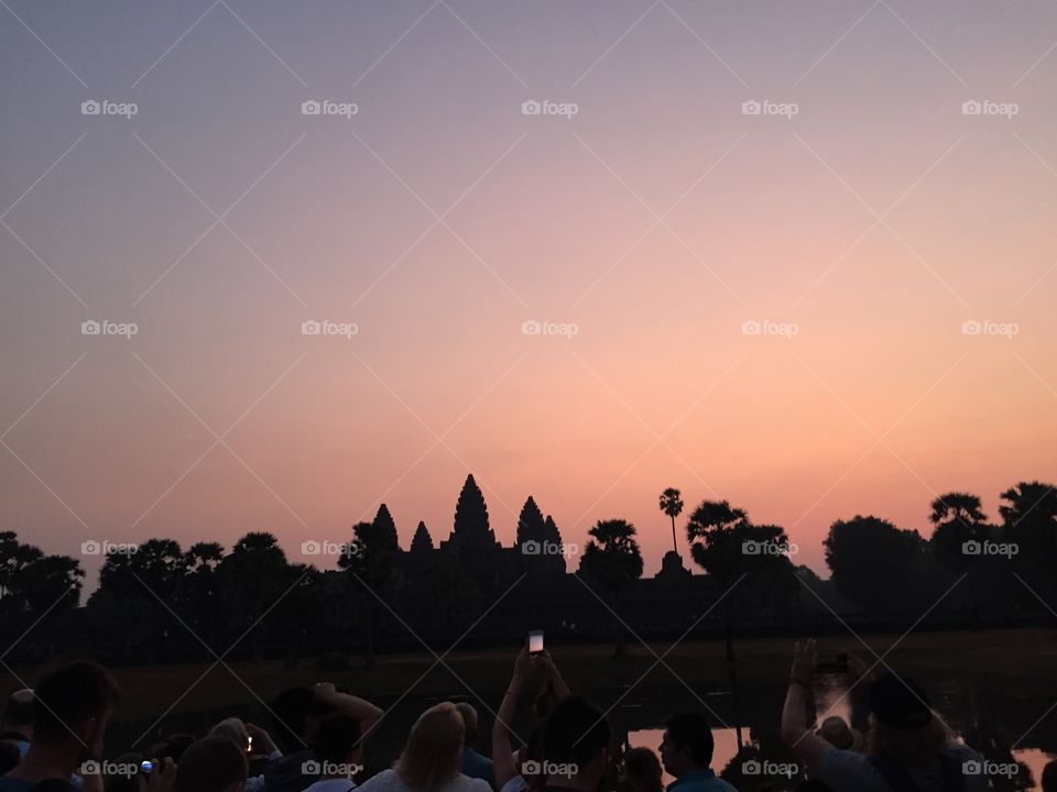 Angkor Wat temple in Cambodia with vibrant sunrise of pink and purple. People in foreground watching the sunrise. Silhouette of temple shown.