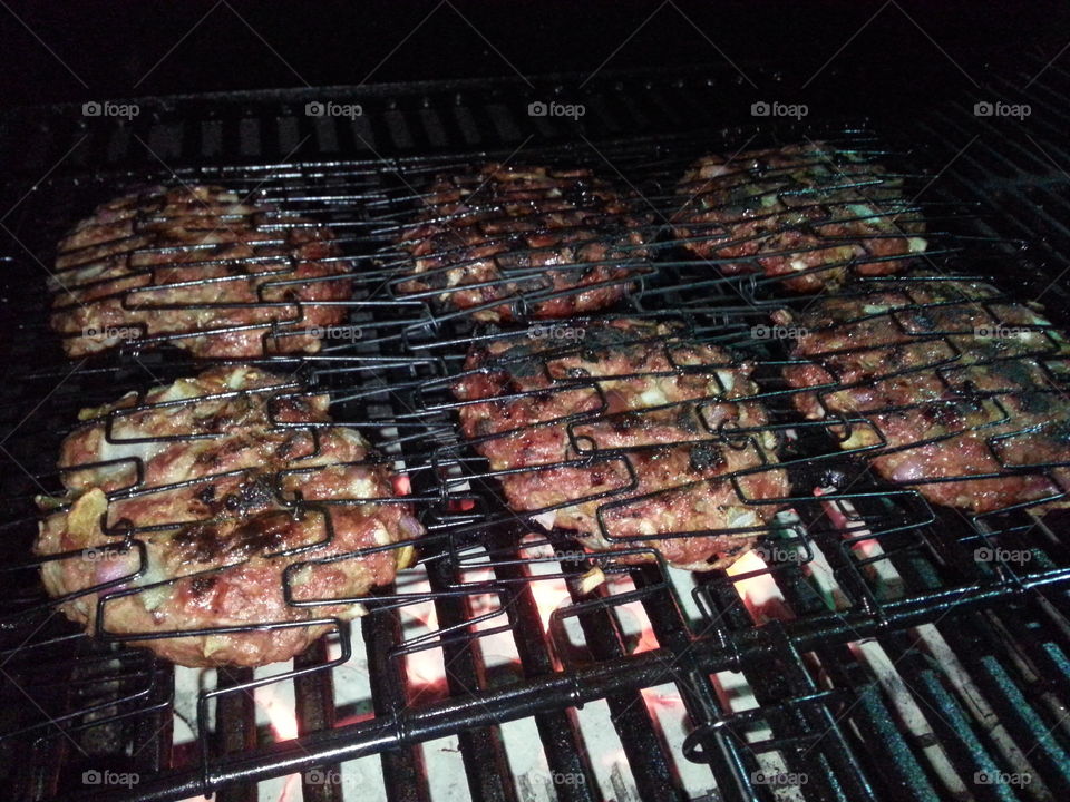Grilling at night. Six burger patties in a grill cage sizzling over a high charcoal flame.