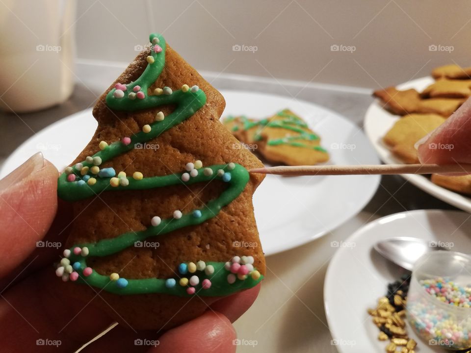 A person is decorating spruce shape ginger biscuits with green sugary coating and multicoloured nonpareils using a wooden stick. In the background some plain and ready cookies on the white plates.