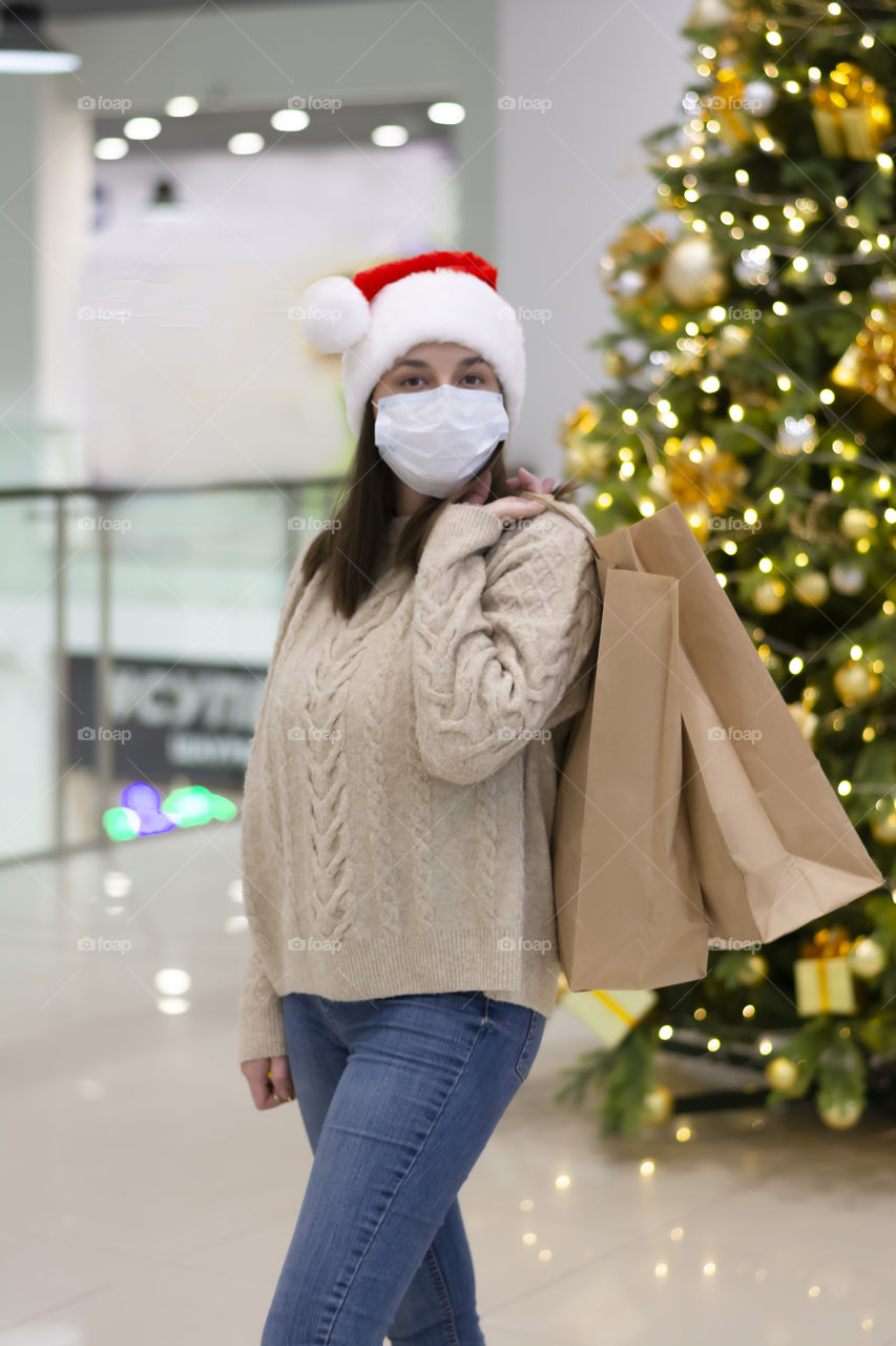Holiday season shopping woman in protective medical mask, christmas hat and knitted sweater in mall with empty craft bags near christmas tree.  Concept: holiday shopping during the coronavirus epidemic a woman wearing a medical mask