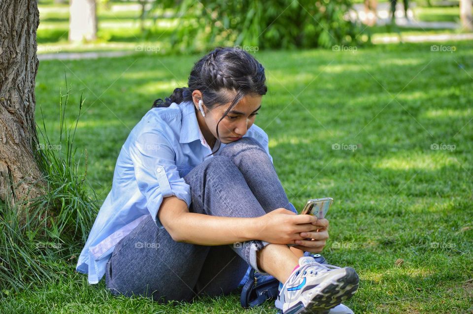 a girl in jeans and a shirt sits on a lawn in a park with a phone in her hands