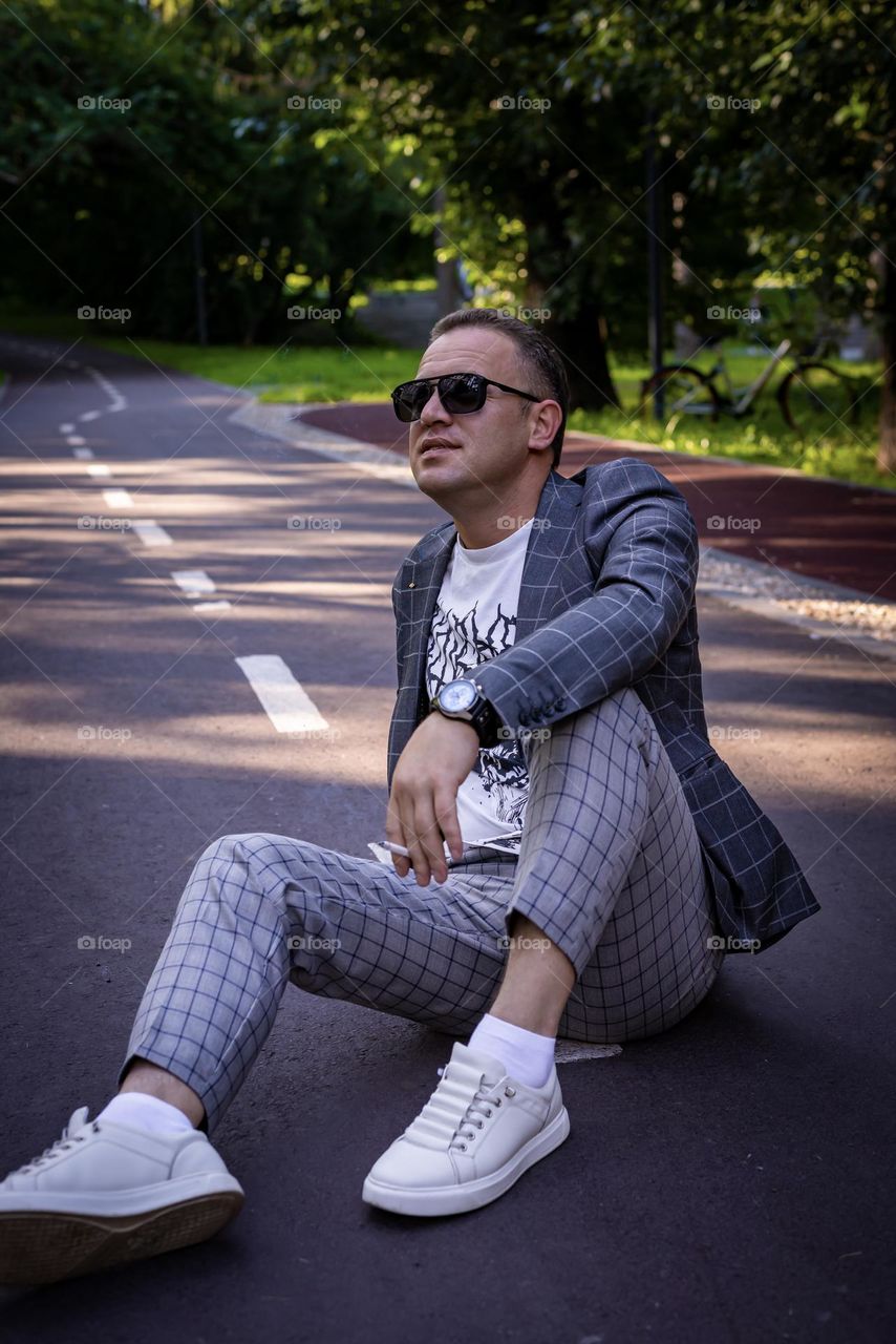 young man on a walk in the city park, summer. Life style portrait sits on the road laughing, in white sneakers and a suit in a cage