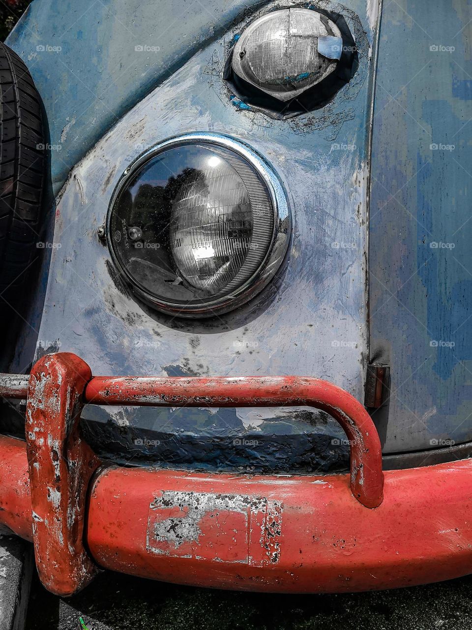 Closeup of and old beat up rusty Volkswagen bus showcasing its red bumper with pealing paint, old tire, headlight, turn signal 