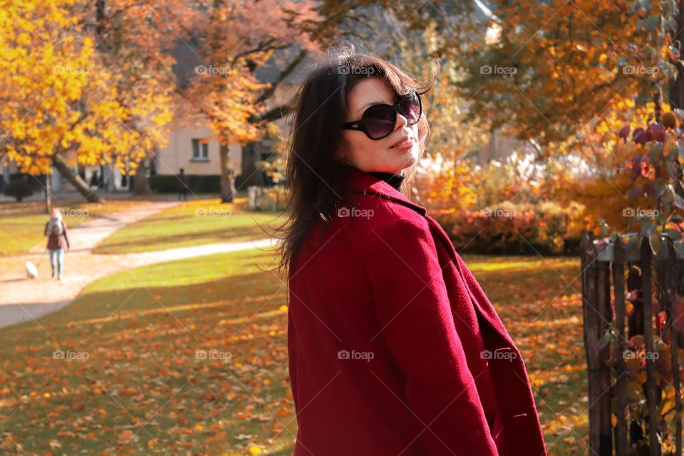 Close-up side view portrait of a beautiful young caucasian brunette girl in a red coat and sunglasses standing in a colorful autumn city park in Brussels Belgium. Concept autumn walk.