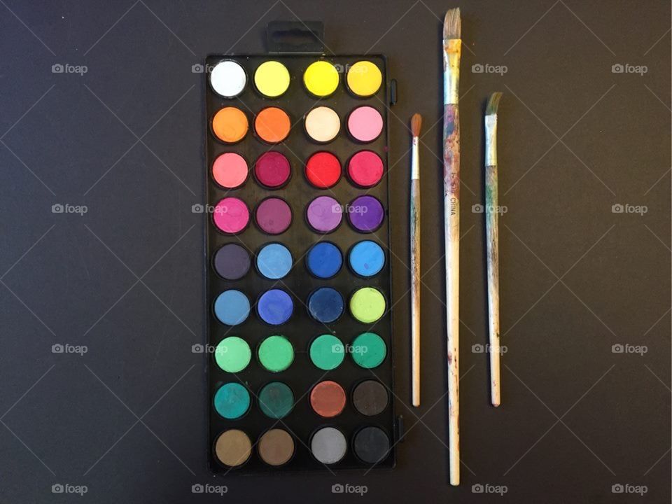 A bright, colorful watercolor set with various paint brushes lined up next to it on a black background 