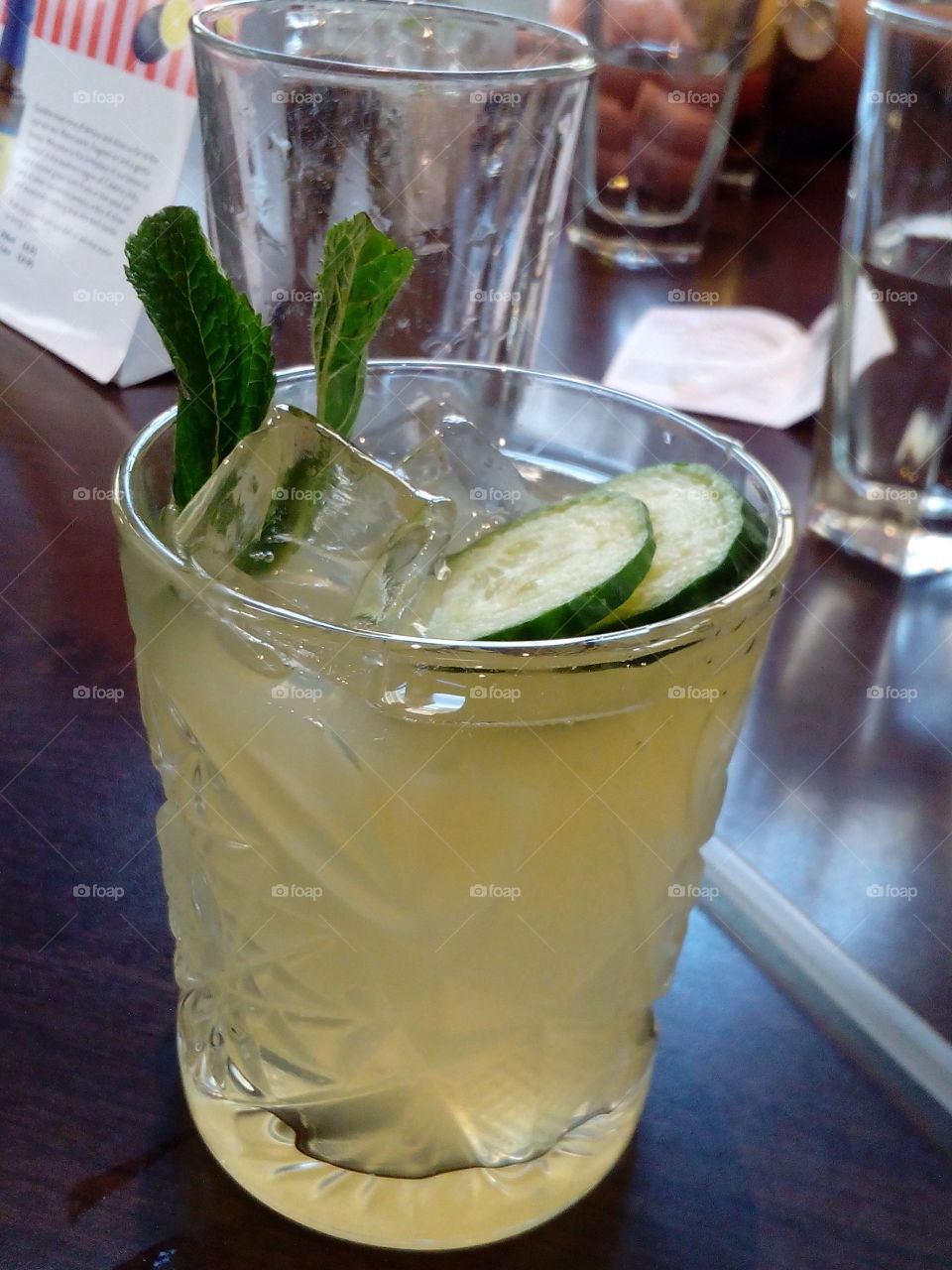 A cocktail on the brown table. The glass is full of the ice cubes, two slices of the cucumber, a green leaf and light yellow liquid. Three glasses behind and a box.