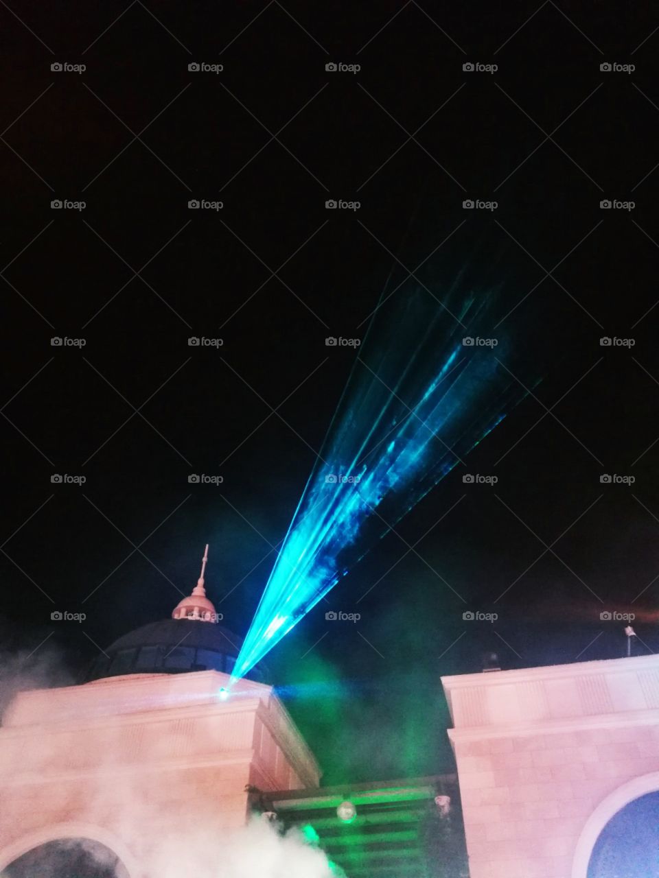 An artificial blue laser light in a festival show with steam below, nice part of building and dark night sky.