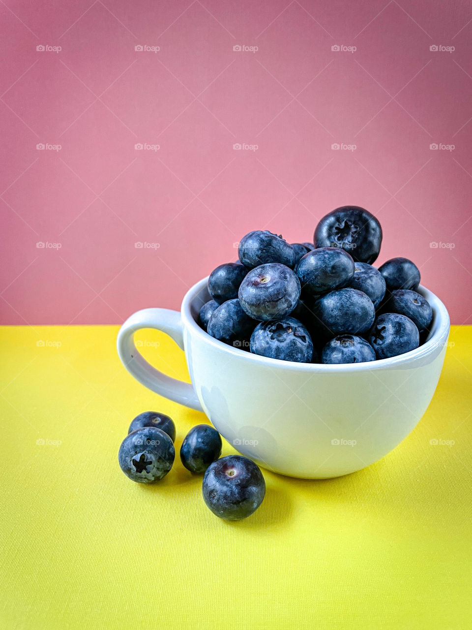 Healthy blueberries in a white cup with a bright yellow and pink background. 