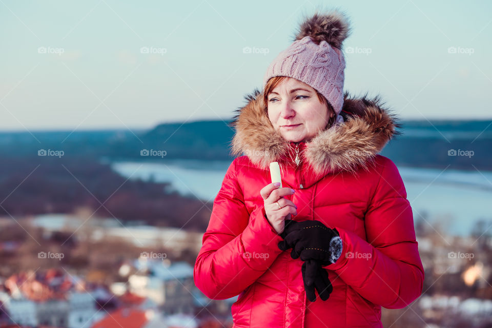 Woman applying lip balsam while walk on a wintery day. Wearing red coat and cap. Town in the background