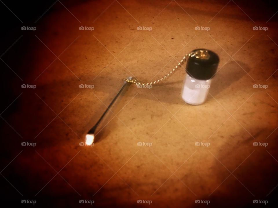 A close up still life of a small cocaine vial with a spoon attached by a chain