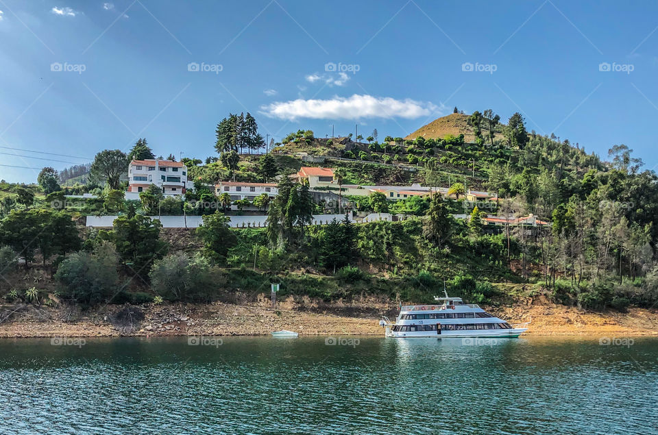 A pleasure boat is docked at a river beach, behind there is a few villas on the hill that leads to the river