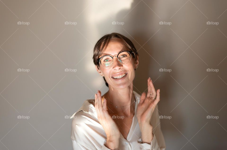 A cheerful young woman with a happy expression is standing against gray wall with hands on her face