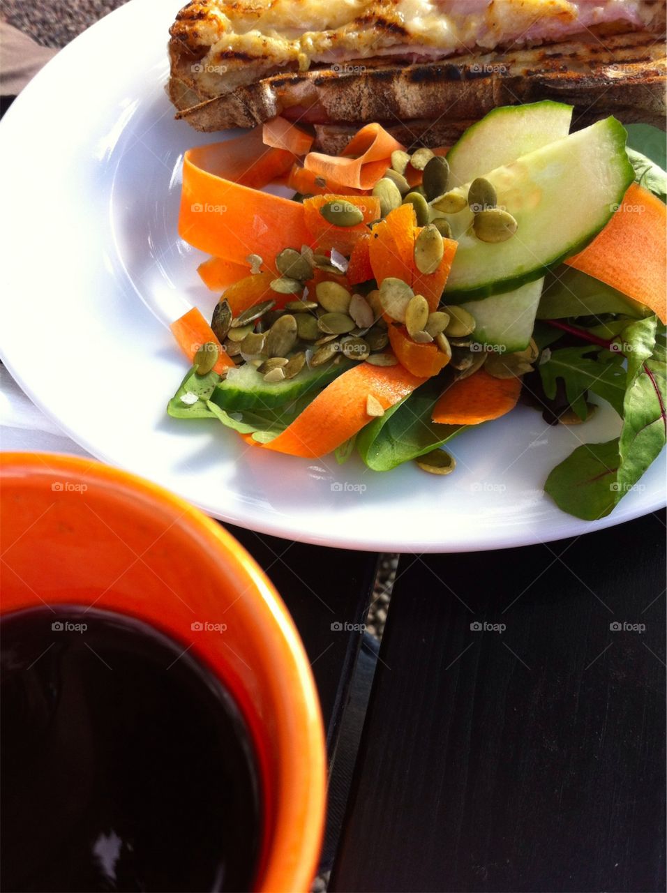 Plate with grilled sandwich and vegetables and a mug with black coffee on a table outdoors.