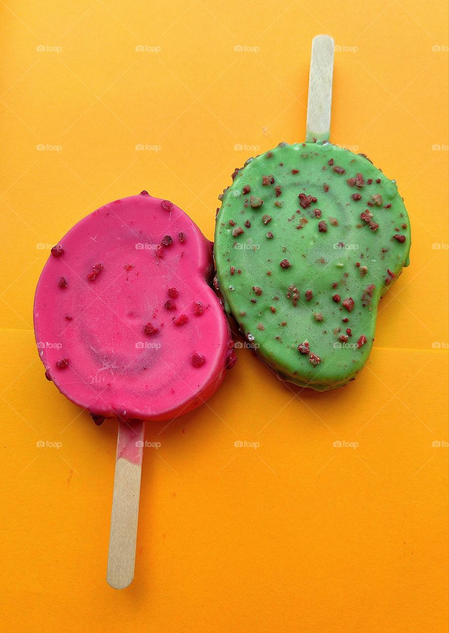 Summer.  Time to cool off.  On an orange background are two ice creams: red raspberry with raspberry pieces and green kiwi with chocolate pieces.  Contrast of colors and taste