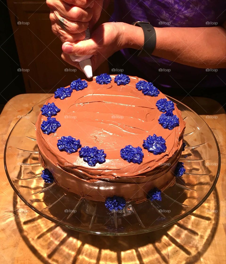 Hands decorating cake with chocolate frosting & a blue design. Glass cake dish & cake are well lit with bright light.