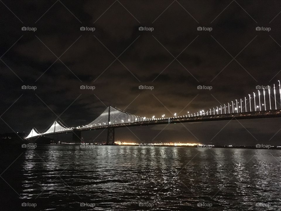 The landmark Bay Bridge in San Francisco lit up at night with light reflecting on the water of the bay below