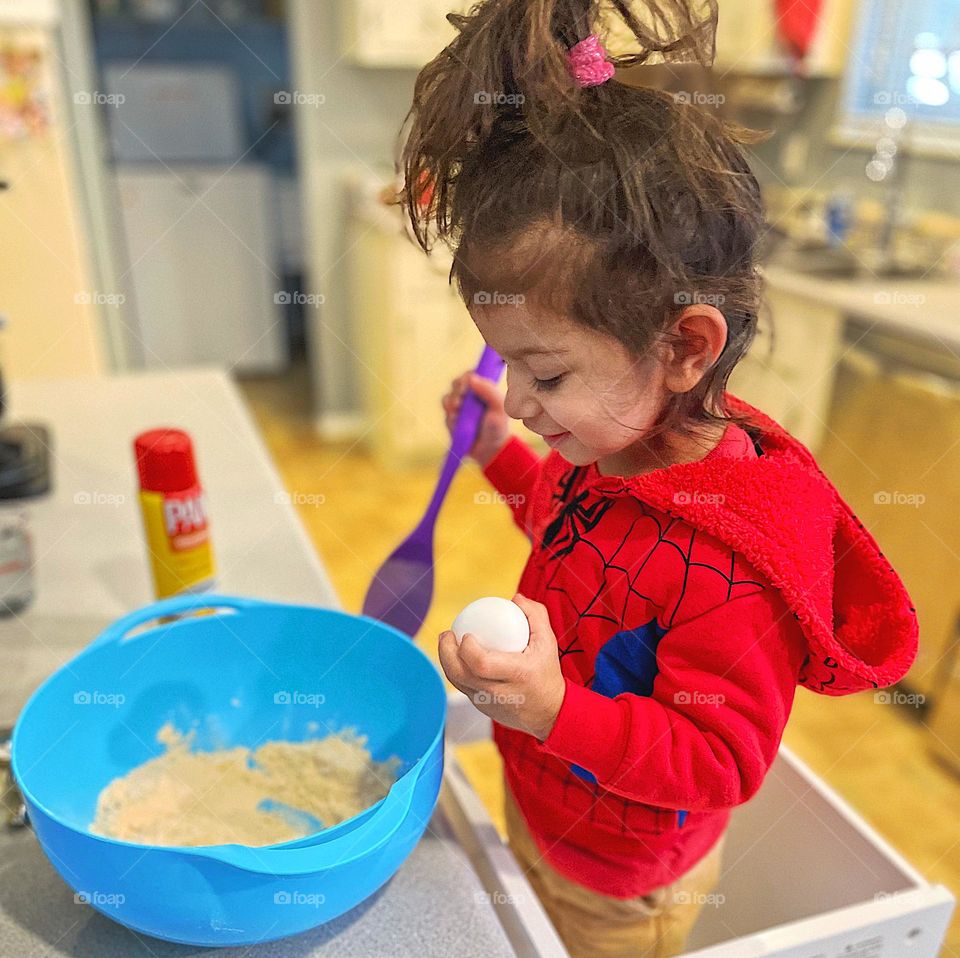 Toddler creating in the kitchen, toddler helps mommy in the kitchen, cooking with toddlers, toddler girl holding egg and smiling, little girl helps cook a creation in the kitchen, mommy’s little helper, eating something you create, creative toddler