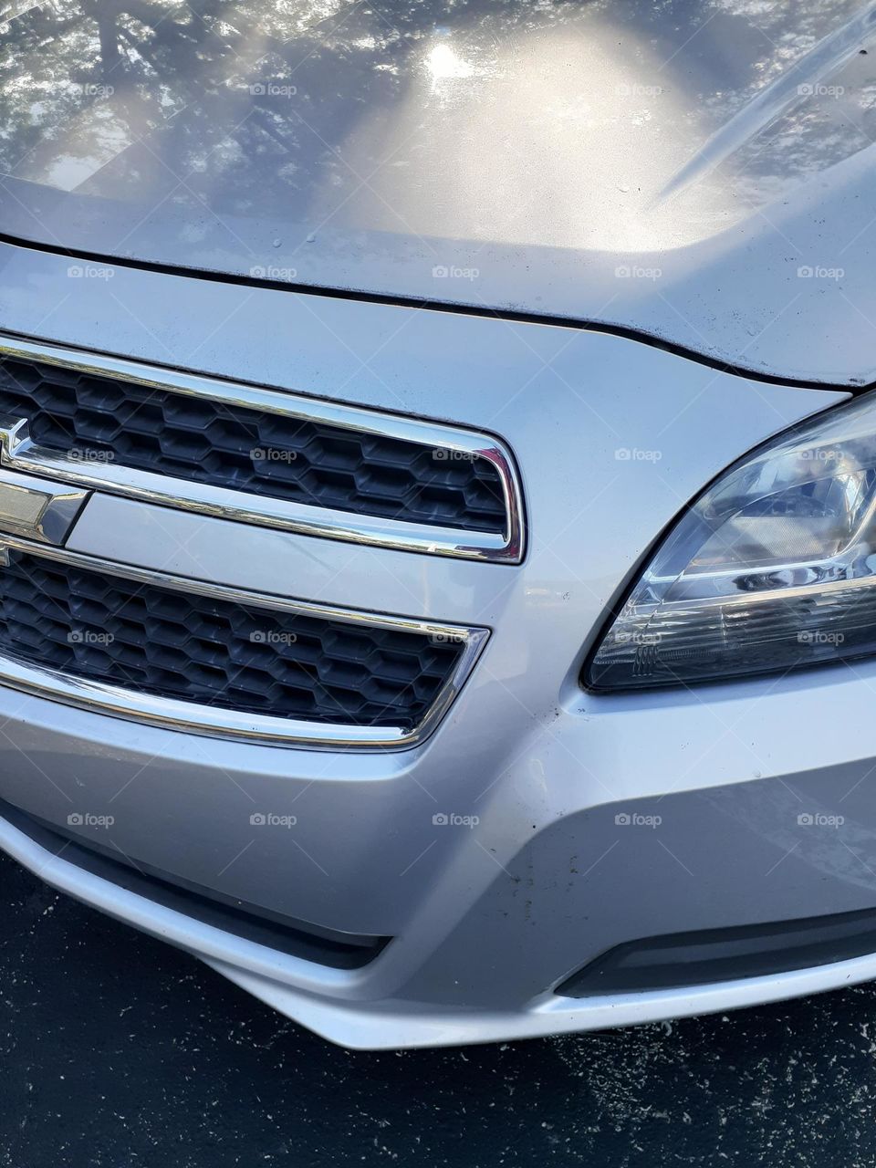 The front left side of a beautiful, sleek, silver 2013 Chevy Malibu. It a sturdy car and a comfortable ride.