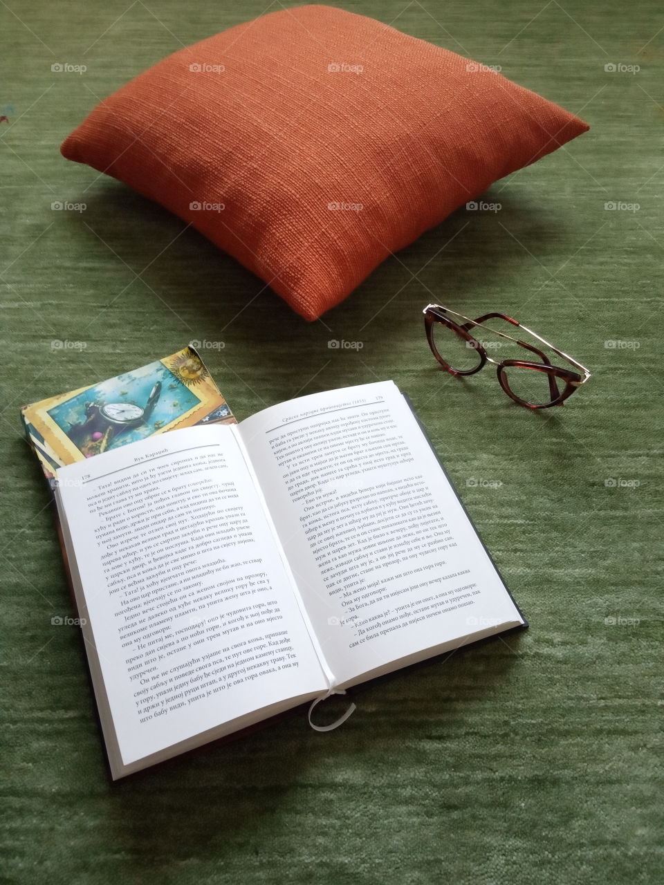 Urban living composition of orange pillow, books and  glasses on calming green surface.