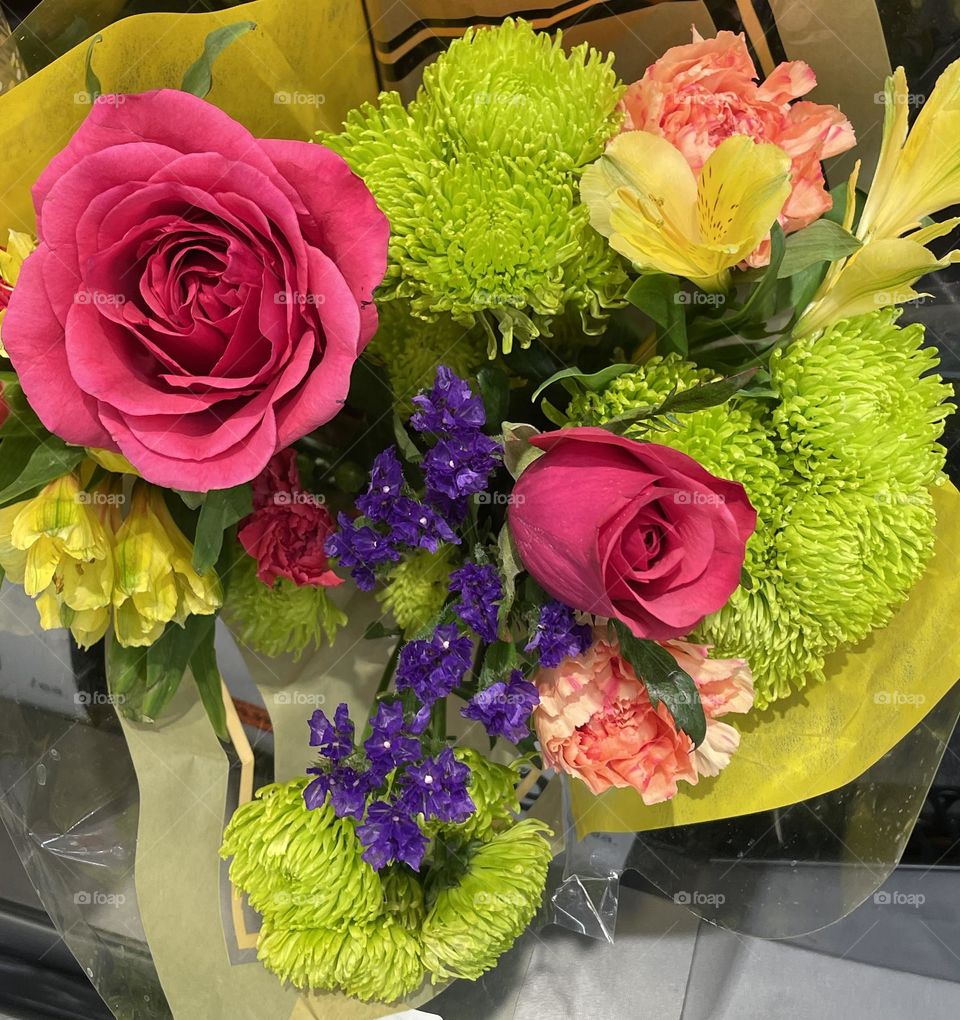A vibrant bouquet bursting with color, featuring lush green mums, bright pink roses, warm peach carnations, and sunny yellow lilies. Sprigs of tiny purple flowers delicately weave through the arrangement, adding a touch of contrast and depth.