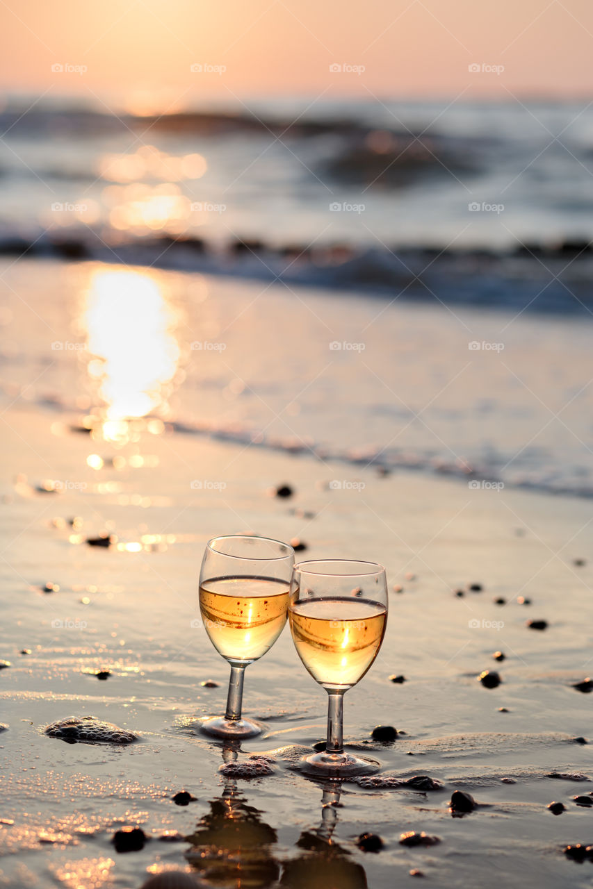 Two wine glasses with white wine standing on sand on beach in the foreground. Blurred sea waves in the background