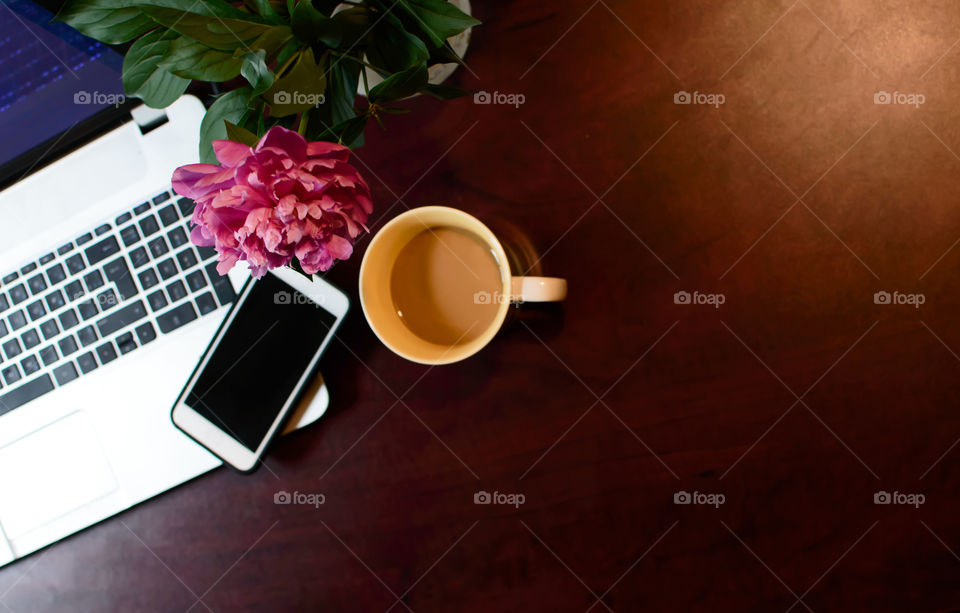 High angle view of computer laptop, mobile phone, technology on desk with peony flowers in vase and cup of coffee conceptual working and business paper free minimalistic wood desk background 