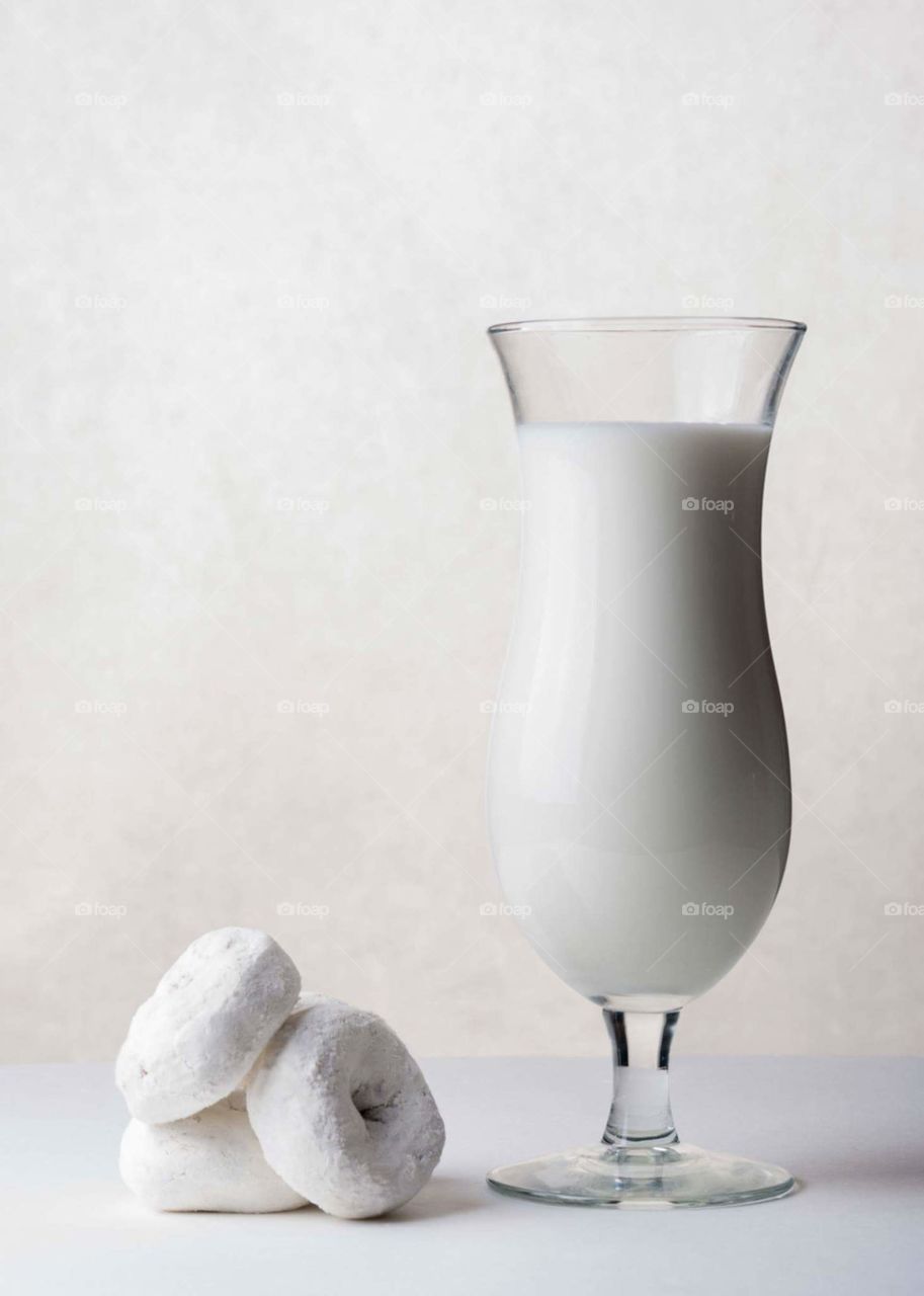 monochrome image of small white powder donuts stacked next to a glass of white milk on a white table in front of a white background