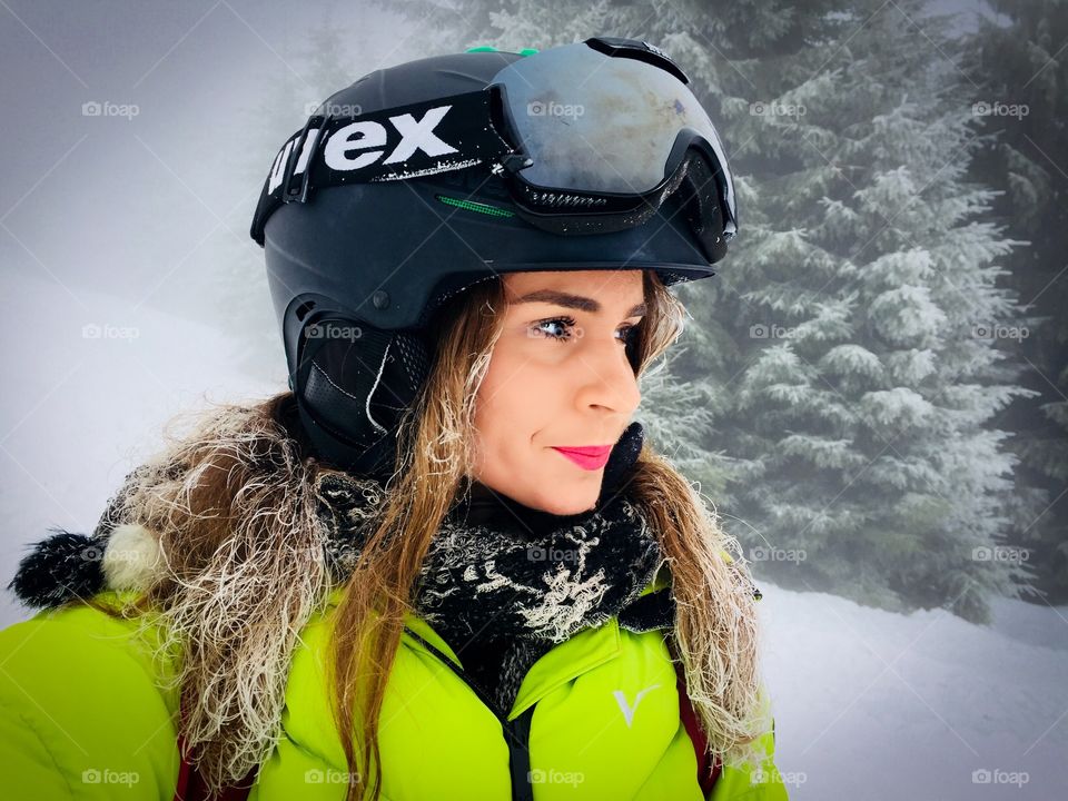Portrait of woman wearing Uvex ski glasses and Uvex ski helmet and holding ski poles with pine cone trees covered in snow in the background
