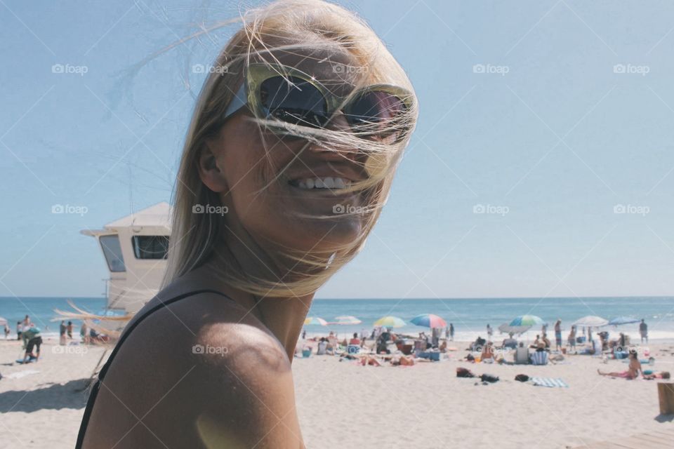 A woman smiles as hair gets whipped in her face from the breeze on a nice beach day. 