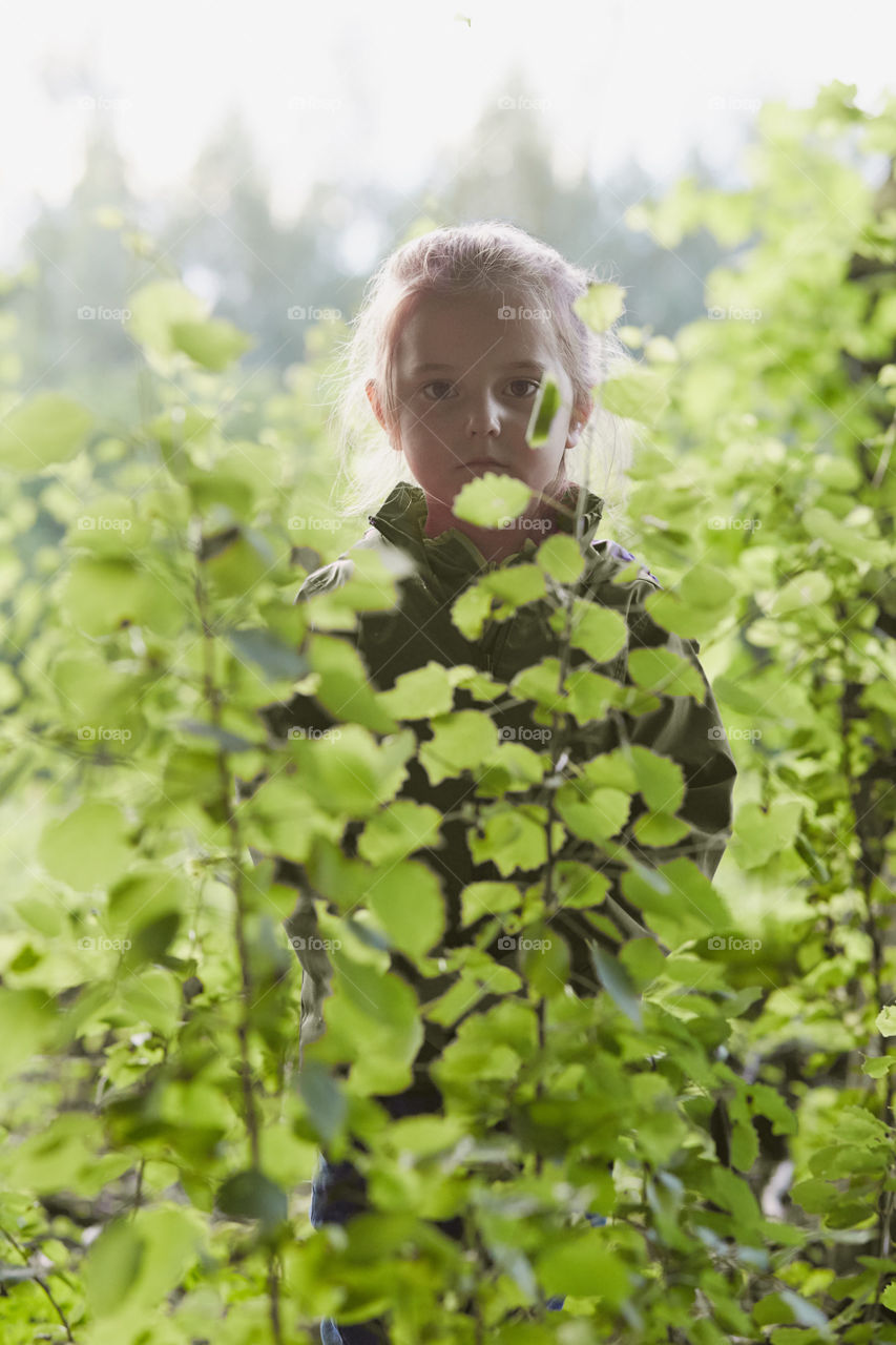 Little girl standing in forest behind the leaves, looking at camera, wearing green clothes. Candid people, real moments, authentic situations