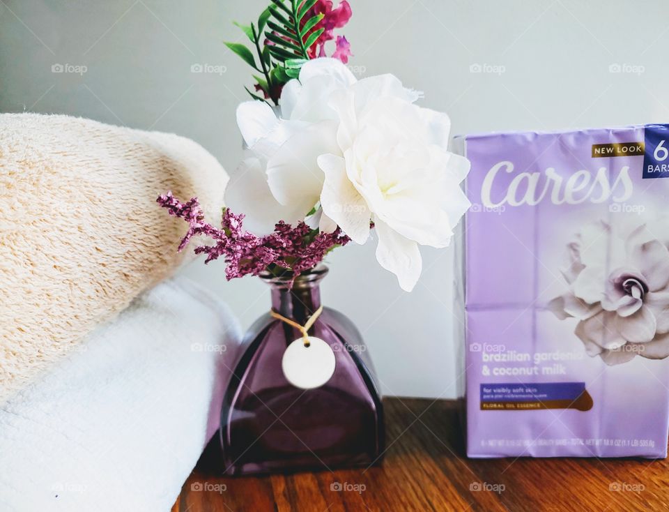The new look Caress brazilian gardenia & coconut milk six pack bar soap accented by a purple vase and white flowers and with a tan and a white towel stacked on the left.