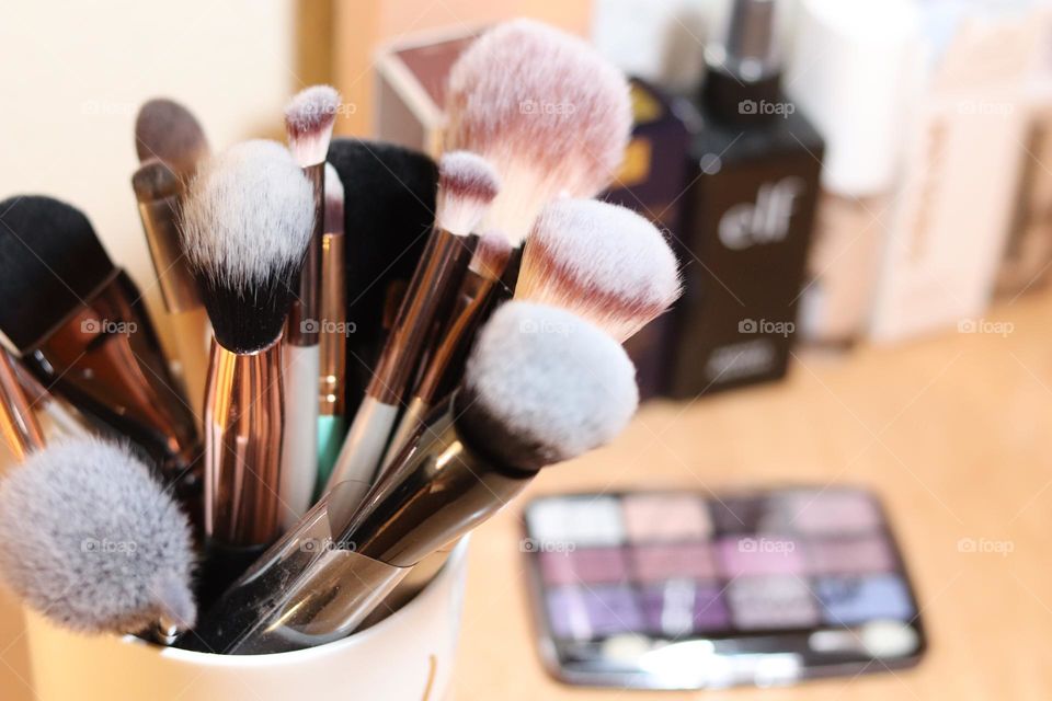 Large collection of makeup brushes in a cup on top of dresser with eyeshadow’s set and other perfume and cosmetics bottles in background 