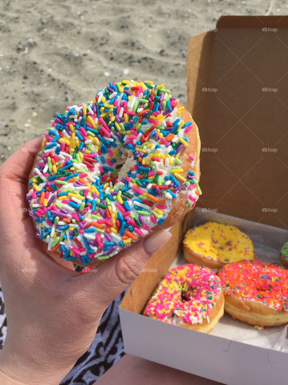 A hand holding a rainbow donut on the beach with a box of donuts in the background 