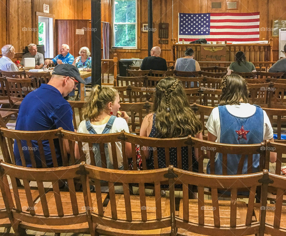 Small town life - enjoying a town BBQ and finding shelter inside the town hall from the rain.  The American flag is prominent in the space. 