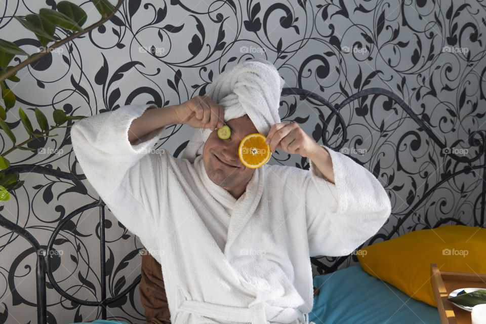 A man and home spa care at home takes care of himself and smears his face with cream, does a manicure at home on a blue background and with oranges and rests.