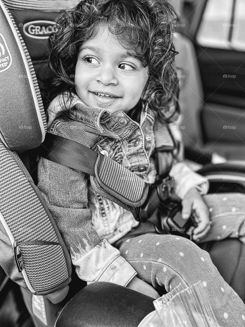 Toddler in car seat looks behind shoulder, toddler looks for strangers, black and white portrait of a toddler girl, monochrome portrait, toddler rides in the car, toddler girl is happy in her car seat, going to the park with mommy, riding in the car 