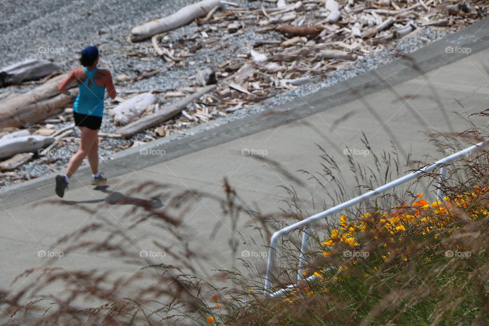 Young woman running on a path by the ocean where grass and poppy flowers are casting their shades under the bright sun