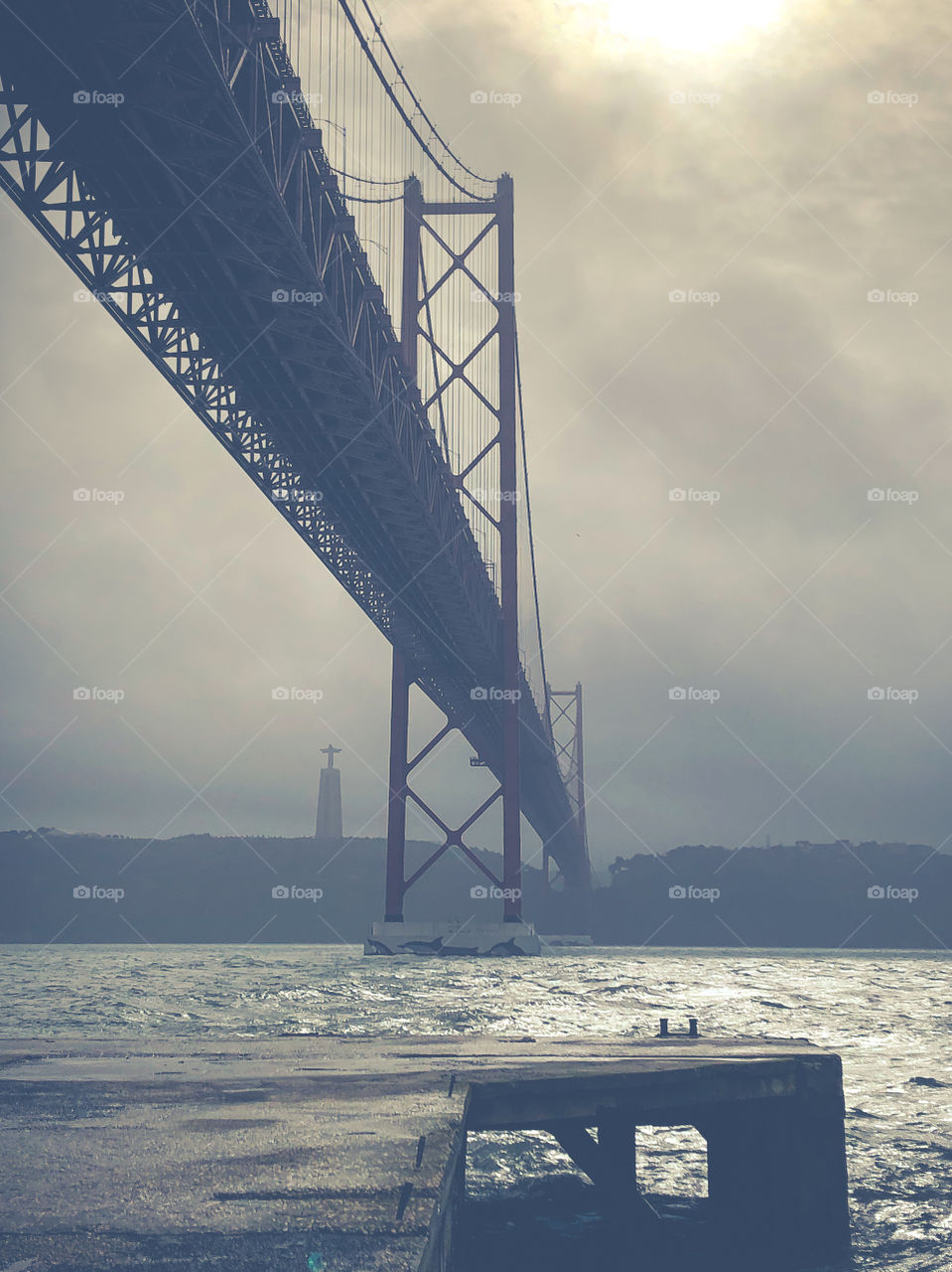 A view from the north bank underneath The Ponte 25 de Abril suspension bridge on a cloudy, misty day in Lisbon