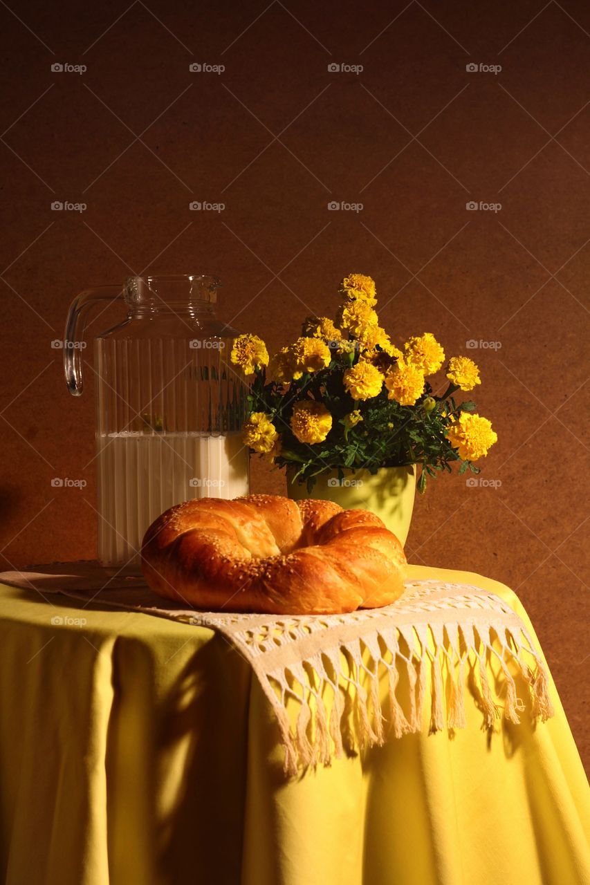 Still life in yellow of a yellow tablecloth, a rushnyka, a glass jug of milk, yellow marigolds in a green vase, and rosy roll of bread